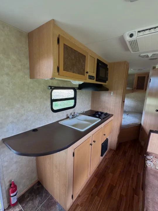 Clean RV rental kitchen with light wood cabinets, dark countertop, double sink, stove, microwave, and bedroom entrance.. kz Coyote 2010