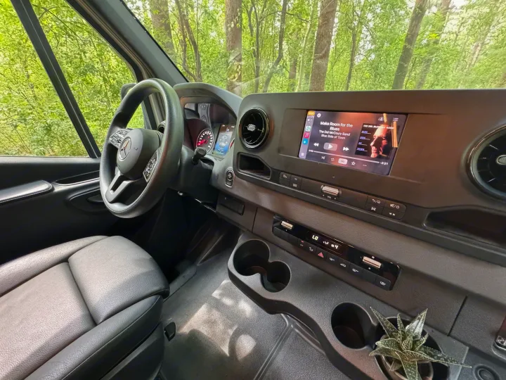 Experience comfort in this RV rental! Modern interior with dashboard, steering wheel, music playing, and a succulent plant by the cups.. Mercedes-Benz Sprinter RV Motorhome Campervan 2022