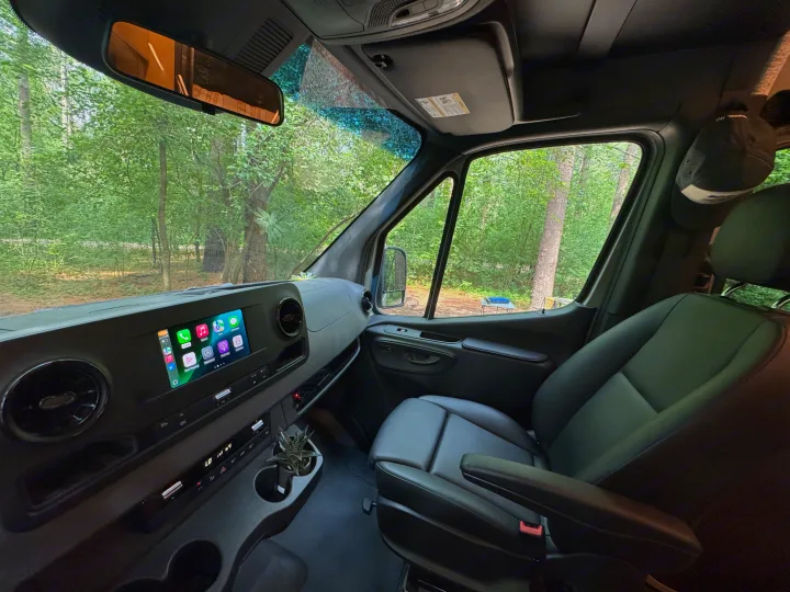 Inside an RV rental van at a forest campsite: dashboard with digital display, leather passenger seat, and outdoor table.. Mercedes-Benz Sprinter RV Motorhome Campervan 2022