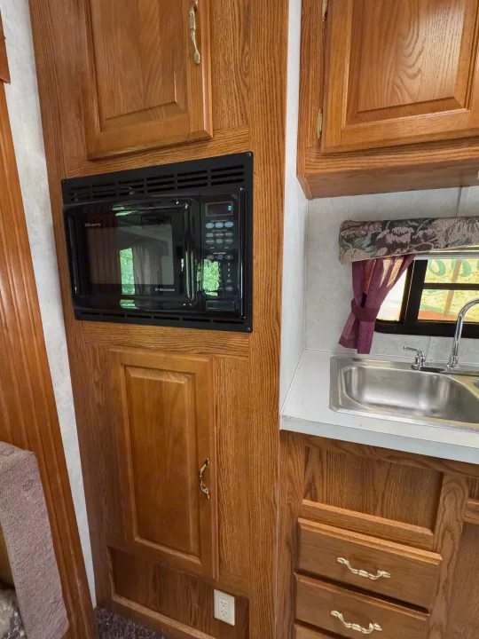 RV rental kitchen features: wood cabinets, Dometic microwave, sink, and window with curtain.. Forest River Wildwood 2000