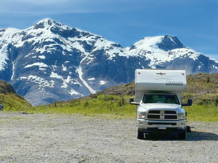 White Lance RV rental, a truck camper, on a gravel lot with majestic, snow-capped mountains under a clear blue sky.. Dodge RAM 3500HD 2010