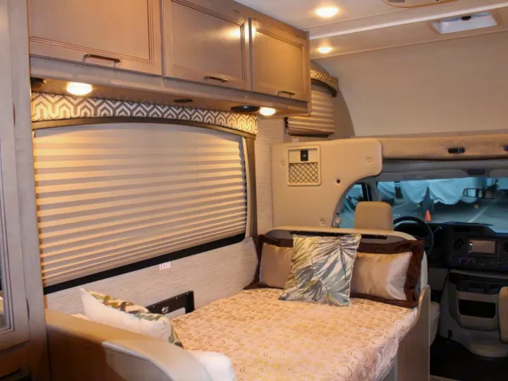 Cozy rv rental interior: a comfortable bed with patterned bedding, overhead cabinets, and front driving area.. Thor Chateau 2022