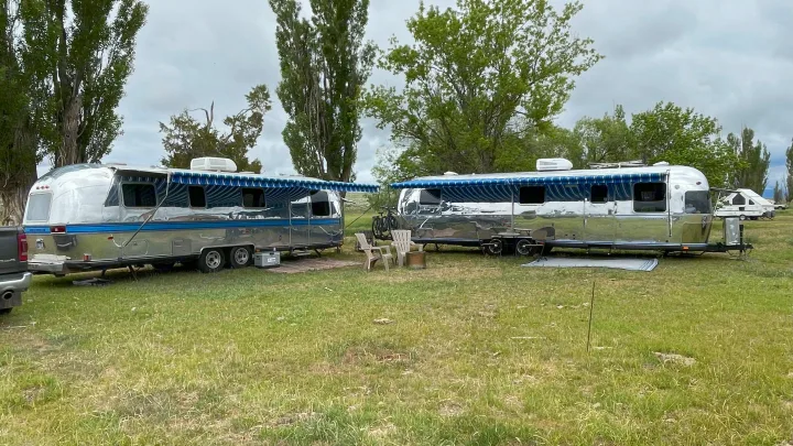 Two shiny Airstream RV rental travel trailers with extended blue awnings, set up for camping on a grassy field.. Airstream Excella 1981