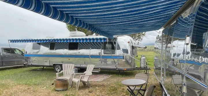 Two polished Airstream travel trailers with blue awnings at a serene RV rental campsite, perfect for outdoor relaxation.. Airstream Excella 1981