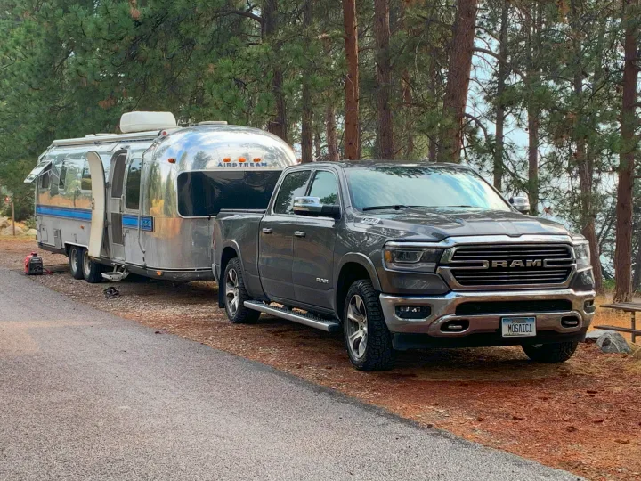 A grey Ram pickup truck tows a polished Airstream travel trailer, parked in a pine forest, perfect for an RV rental adventure.. Airstream Excella 1981