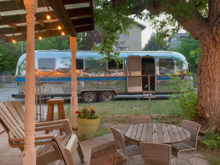 An Airstream RV rental is parked beyond a cozy patio with outdoor seating and string lights, surrounded by trees at dusk.. Airstream Excella 1981