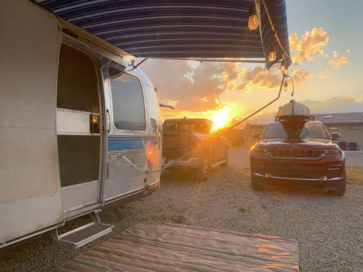 Golden sunset lights an RV rental setup: an Airstream, truck, and Jeep with canoe on a gravel lot.. Airstream Excella 1981