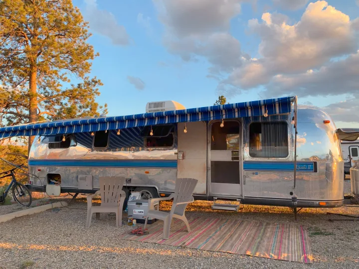 Shiny Airstream rv rental at a cozy gravel campsite. Blue awning, chairs, rug, & string lights invite outdoor relaxation.. Airstream Excella 1981