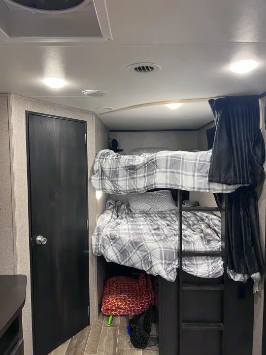 RV rental interior: two cozy bunk beds with plaid bedding. A dark door, ceiling lights, and vents define this compact space.. Jayco Jay Feather 2021