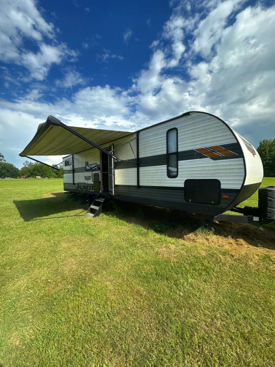 RV rental travel trailer set up with awning in a grassy field under a cloudy sky.. Forest River Wildwood 2021