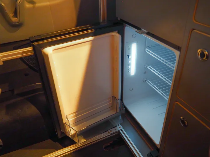 Inside a clean, brightly lit compact refrigerator in an RV rental, showing wire shelves, a door bin, and control panel.. Ford Transit 2019