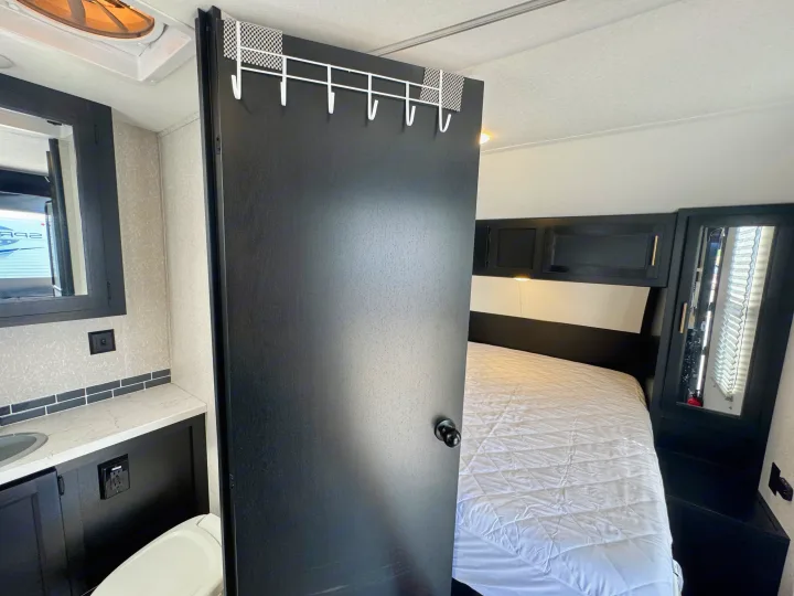 Inside a sleek **rv rental**: a modern bathroom area with vanity & toilet, adjacent to a bedroom with a quilted bed.. Forest River Grey Wolf Limited 2022