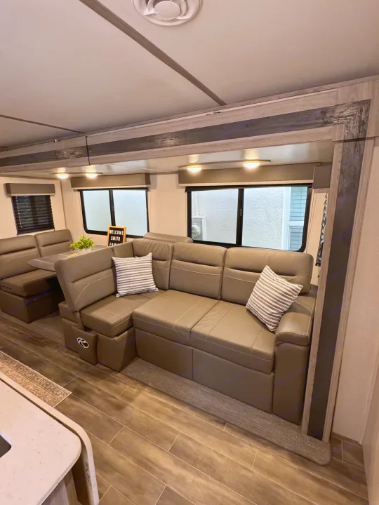Modern RV rental living area featuring a large L-shaped sofa, dinette, windows, and wood-look tile floors.. Keystone RV Passport TT 2022