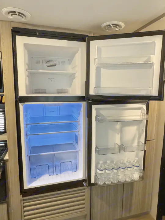 Open Dometic RV rental fridge and freezer, showing interior shelves, frost in freezer, and water bottles.. Keystone RV Passport TT 2022