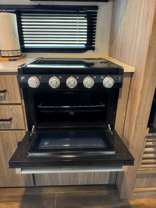 Inside an RV rental: a modern, black gas oven with an open door and stovetop controls. Perfect for travel cooking.. Keystone RV Passport TT 2022