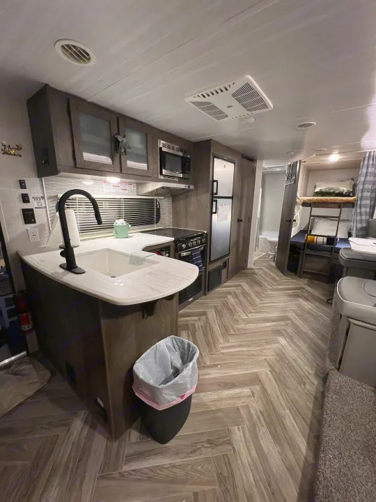 View of a modern rv rental kitchen with white counters, dark cabinets, a fridge, and a glimpse of bunk beds.. Forest River Wildwood X-Lite 2022