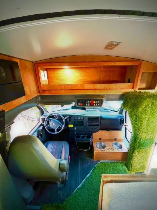 View of a custom rv rental interior from passenger seat: driver's area, wood storage, pet bowls on green turf.. Chevrolet Off Grid Skoolie 2012