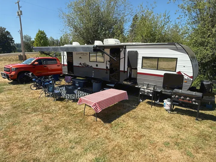 An RV rental travel trailer campsite setup with an orange truck, chairs, outdoor cooking griddle, and picnic table.. Forest River Campsite reserve 26CJ 2025