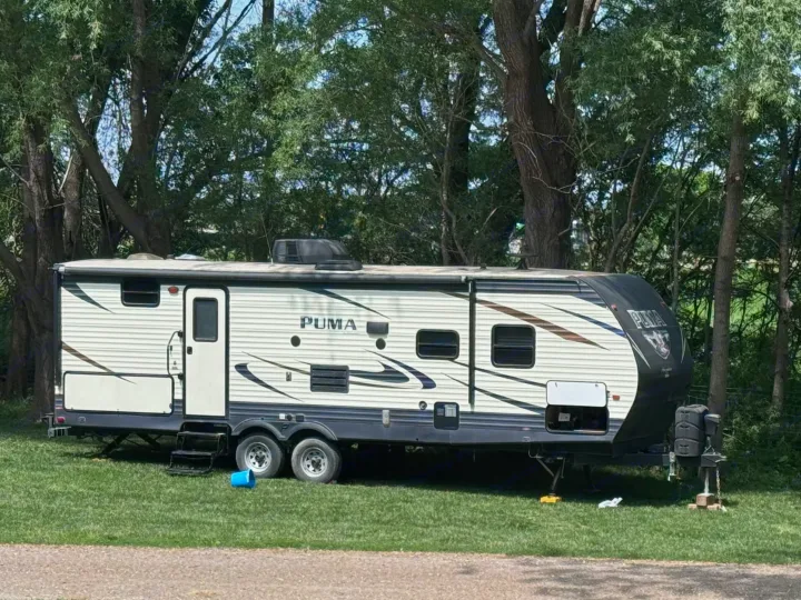 A spacious PUMA travel trailer, ideal for an RV rental, is parked on lush green grass amidst shady trees. Ready for your next adventure.. Forest River Palomino Puma 2018