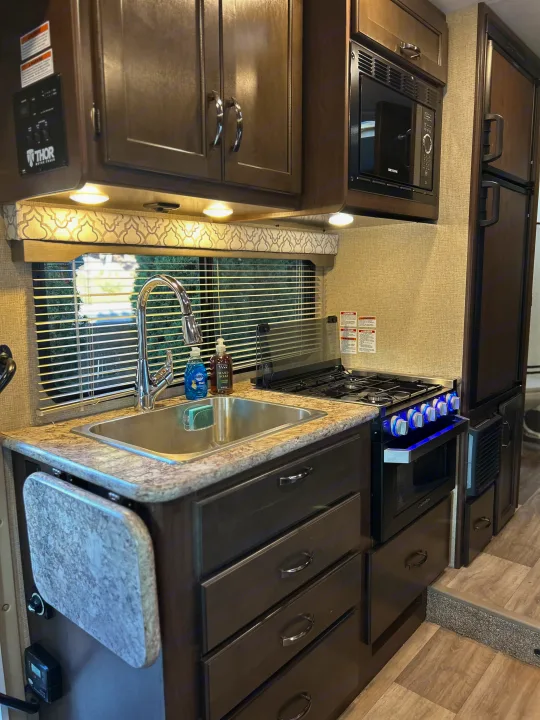 Explore this RV rental kitchen featuring a stainless sink, microwave, gas range with blue-lit knobs, and ample cabinet storage.. Thor Freedom Elite 2019