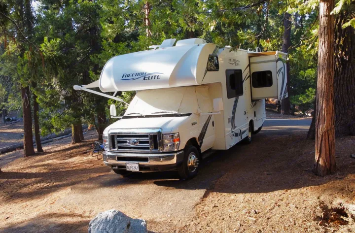 Enjoy the outdoors! This spacious white RV rental motorhome is parked at a sunny, tree-lined campsite, perfect for travel.. Thor Freedom Elite 2019