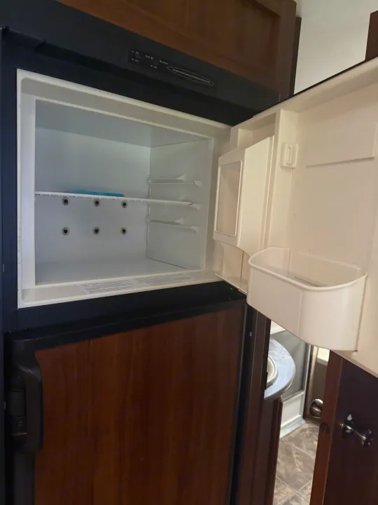 Empty RV rental refrigerator, ready for stocking. Shows open fridge interior with controls and wood cabinetry.. Keystone RV Cougar 2014