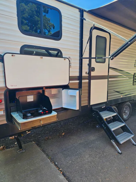 RV rental side view featuring an open outdoor kitchen with a stove and fridge, an extended awning, and entry steps.. Dutchmen Coleman Lantern 2021