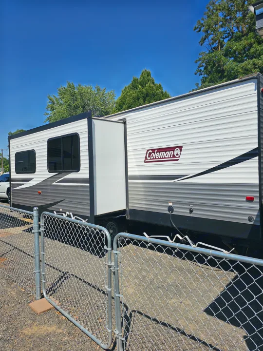 Modern white and gray Coleman travel trailer, slide-out extended, behind a fence. Great for rv rental or camping.. Dutchmen Coleman Lantern 2021