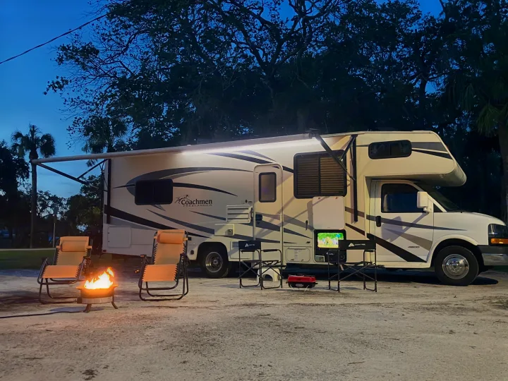 Evening scene with an RV rental, featuring a Coachmen motorhome. A warm fire pit, camping chairs, and outdoor TV create a cozy outdoor retreat.. Forest River Coachmen Freelander 2017
