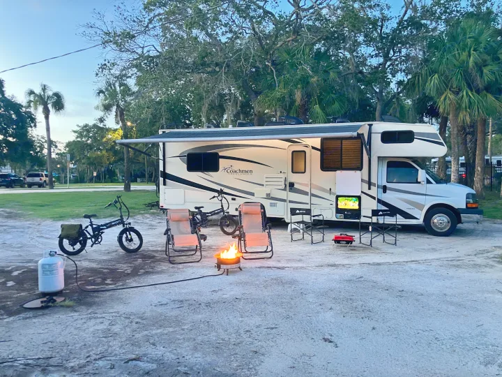 An RV rental is set up for camping with bikes, a fire pit, and outdoor TV, providing a relaxing evening scene among trees.. Forest River Coachmen Freelander 2017