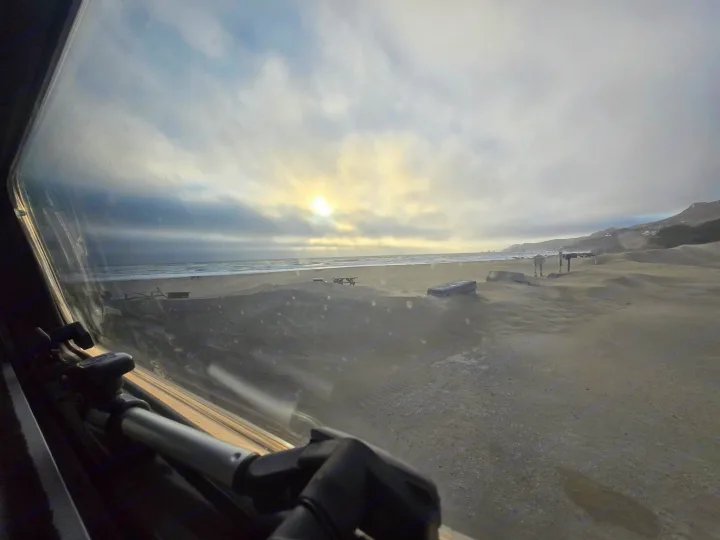 Peeking out our RV rental window to a vast sandy beach, ocean, and cloudy sky. Serene coastal landscape.. Entegra Launch Sprinter High Roof AWD 4x4 Camper Van 2025
