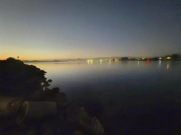 Tranquil water at dusk with glowing distant lights, ideal for an RV rental trip. Dark rocky shore in view.. Entegra Launch Sprinter High Roof AWD 4x4 Camper Van 2025