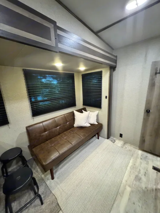 Interior of an RV rental: a brown futon with white pillows, two windows with blinds, light rug, and black stools.. Keystone RV Sprinter Campfire Edition 2020