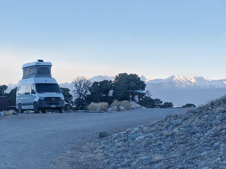 A white RV rental camper van is parked on a dirt road, offering stunning views of snow-capped mountains.. Mercedes-Benz Sprinter 2020