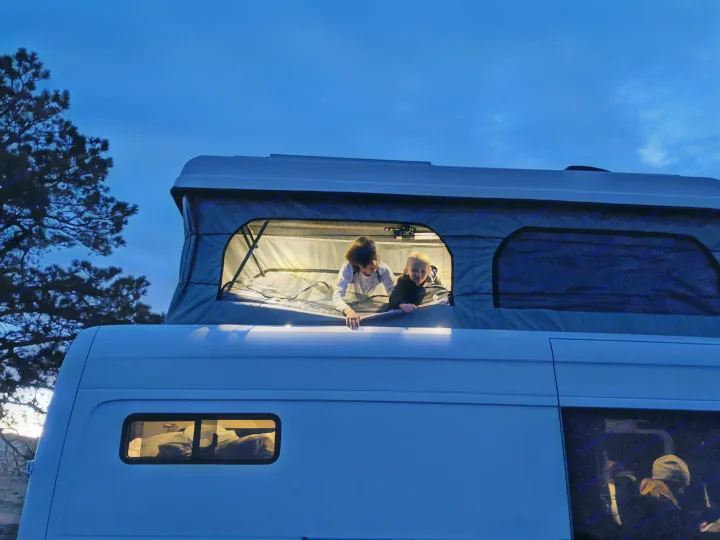 Two kids peer from a lit pop-top camper, an **RV rental** perfect for evening adventures.. Mercedes-Benz Sprinter 2020