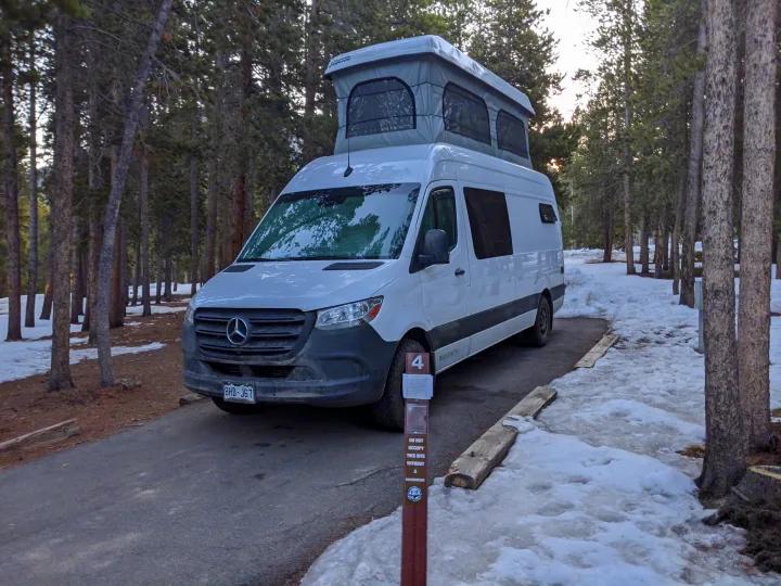 This white pop-top camper van, an RV rental, is parked at snowy campsite #4 in a forest.. Mercedes-Benz Sprinter 2020