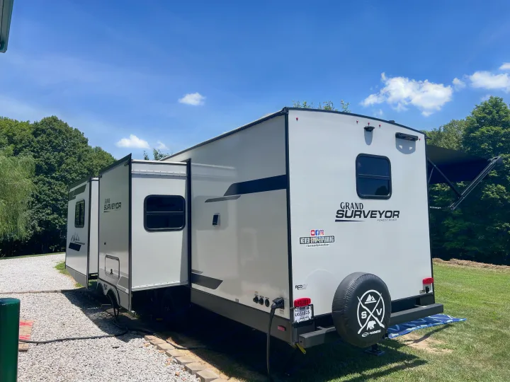 A white Forest River Grand Surveyor **RV rental**, with extended slide-outs and awning, parked at a sunny outdoor campsite.. Forest River Grand Surveyor 2024