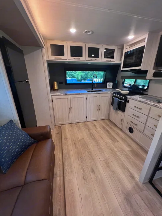 Bright RV rental interior: modern kitchen with light wood cabinets, dark counters, full appliances, and a comfy brown sofa.. Forest River Grand Surveyor 2024