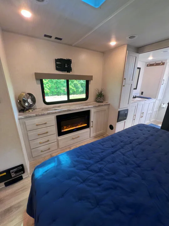 Modern RV rental bedroom featuring a plush blue bed, electric fireplace, and large window with nature views.. Forest River Grand Surveyor 2024
