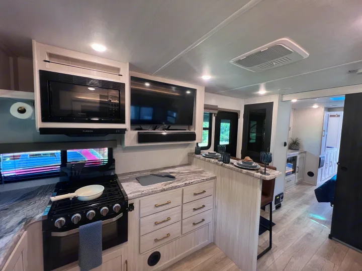 Modern RV rental interior with a bright kitchen (stove, microwave, TV), stone counters, and a dining bar set for meals.. Forest River Grand Surveyor 2024