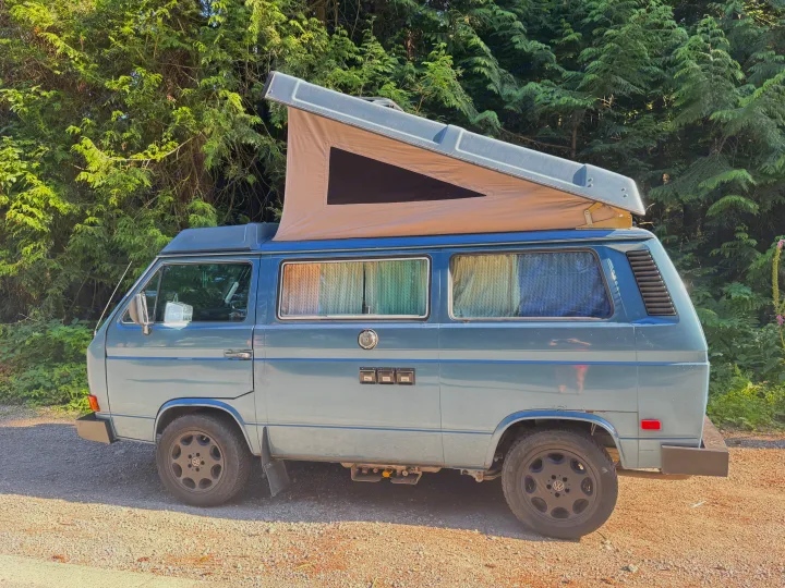 A blue VW pop-top camper van, ideal for RV rental adventures, is parked on gravel with its roof raised near lush trees.. Volkswagen T3 Westfalia 1987