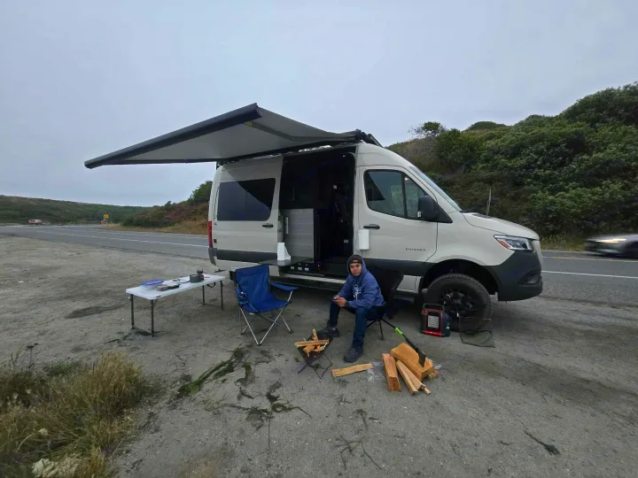 Solo camper enjoying an **rv rental** trip, fireside next to a parked camper van with an awning extended by the road.. Entegra Launch Sprinter High Roof AWD 4x4 Camper Van 2025