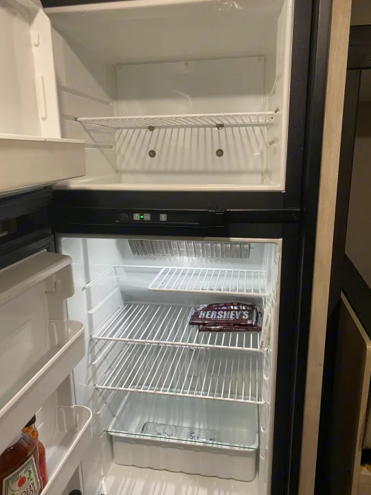 Open RV rental fridge with an empty freezer, Hershey's chocolate, ketchup, and a bottle inside.. KZ Escape 2021