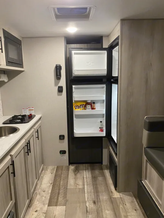 A brightly lit RV rental interior shows a compact kitchen with a sink, stovetop, microwave, and an open refrigerator stocked with basics.. KZ Escape 2021