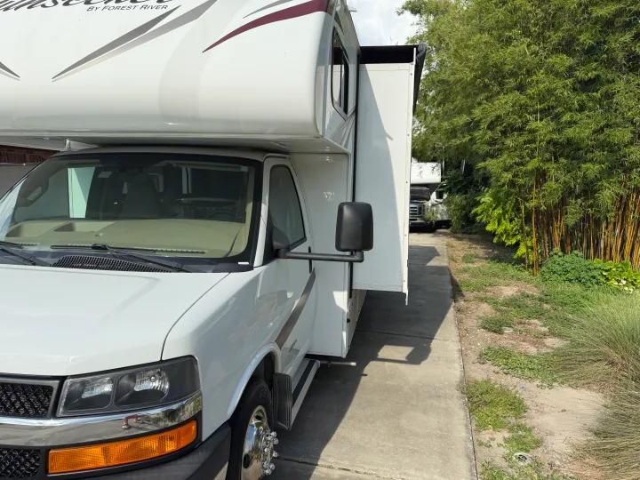 A white Sunseeker RV rental with an extended slide-out, parked on a concrete driveway alongside dense bamboo. Another RV is behind it.. Forest River Sunseeker 2017