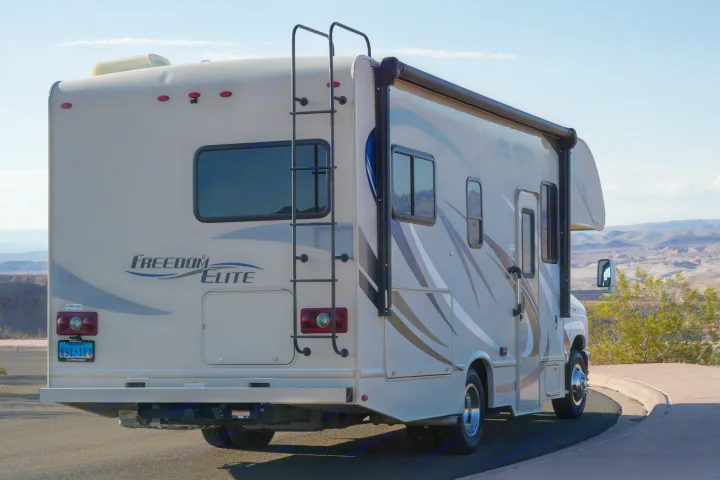 A beige Freedom Elite RV rental, seen from the rear, parked on asphalt with a ladder, against a clear sky and desert mountains.. Thor Freedom Elite 2018