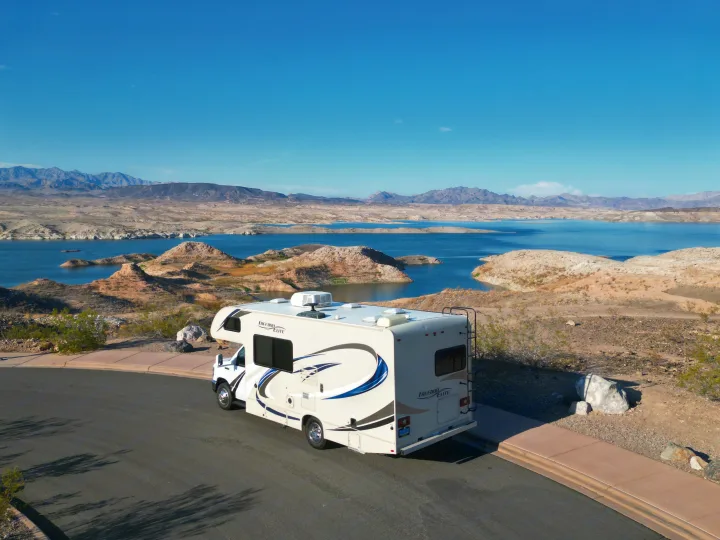 RV rental overlooks a stunning blue lake with exposed shores and distant mountains from a paved road.. Thor Freedom Elite 2018