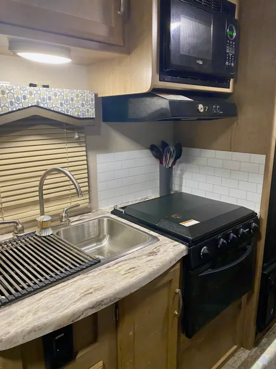 Inside an RV rental kitchen: sink, stove, microwave, range hood, and white subway tile backsplash over light wood cabinets.. Winnebago Minnie Winnie 2020