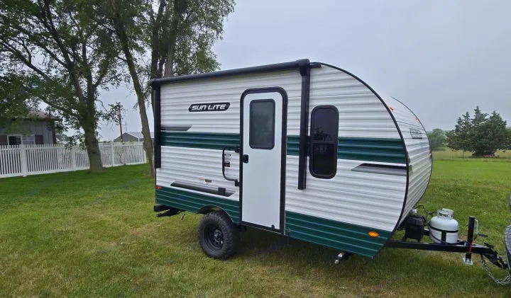 Compact Sun Lite travel trailer, ideal for an RV rental adventure, parked on grass with trees & fence background. Ready for camping!. Sunset Park RV Sun Lite LTD 13MJ 2025