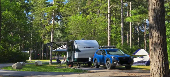 An RV rental travel trailer and blue SUV set up at a vibrant forest campsite with a tent and chairs.. Sunset Park RV Sun Lite LTD 13MJ 2025
